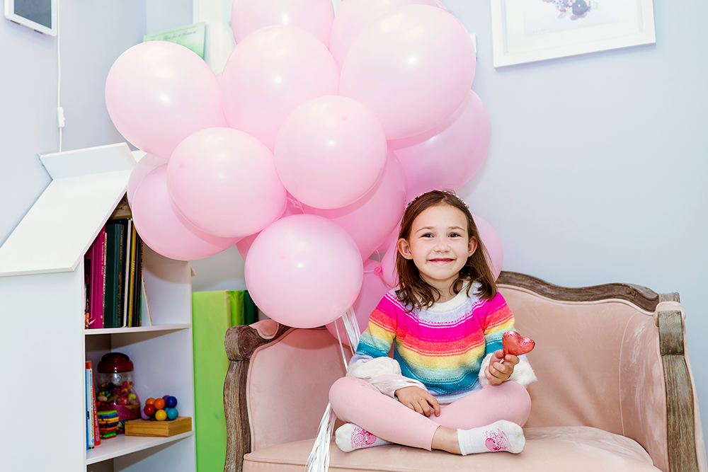 A smiling child, sitting on a child-sized pink sofa, holding a bunch of pink balloons and a shiny heart candy. She is wearing a rainbow striped sweater.