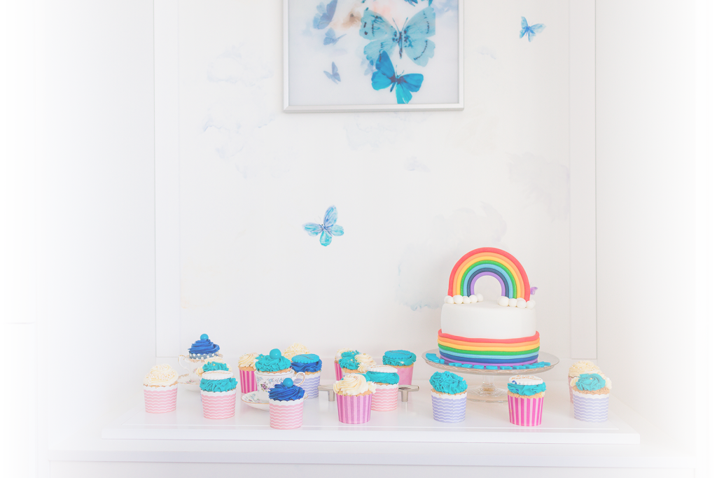 A sideboard topped by colourful cupcakes and a rainbow cake, with a butterflies mural in the background.