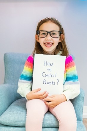A laughing girl, wearing glasses, seated on a child-sized chair, holds a book with a funny handwritten title: "How to Control Parents?". Specialized child therapy services that use a playful approach resonate with kids.