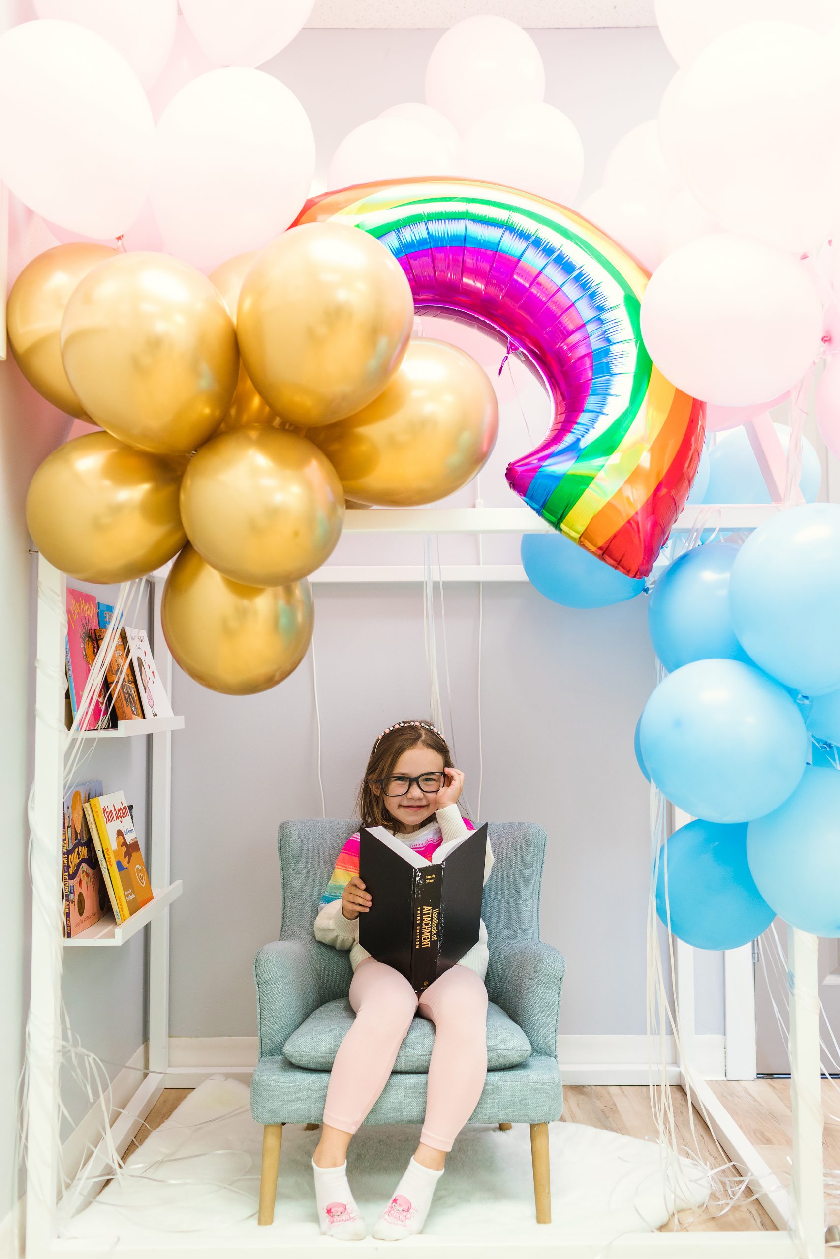 A child smiles as she playfully pretends to read a psychology textbook. She is wearing toy glasses. Gold, pink, blue, and rainbow balloons shine overhead.