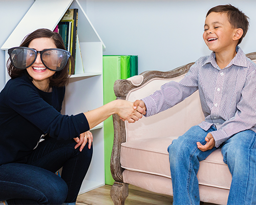 Wearing giant toy glasses, a therapist shakes a child's hand. Both are smiling widely as they meet before a specialized child counselling session!