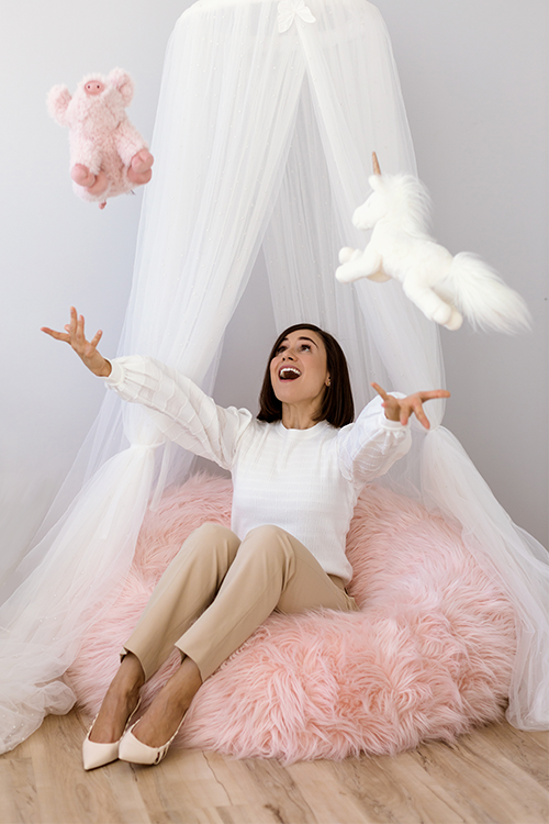 A woman, sitting on a fluffy pink floor pillow, playfully throws a flying toy piggy and unicorn