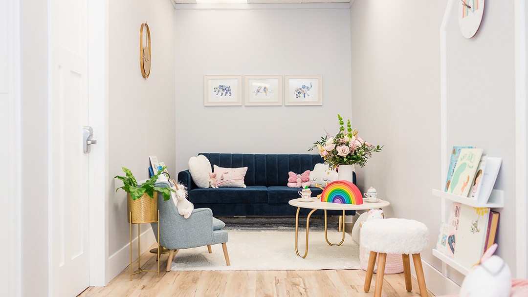 A warm and inviting waiting room. A large navy blue couch with plush toy cushions in the background. Child-sized comfy chair and a fluffy seat on each side. Toys, books placed at child's eye level.