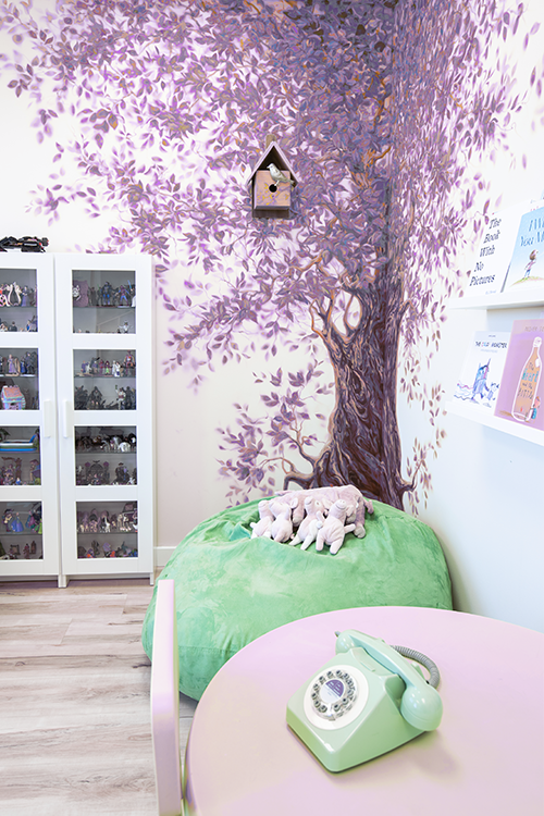 A playroom with a purple tree mural, toy piggies with their Mom on a green floor pillow, and a pink table with a green vintage phone in the foreground.