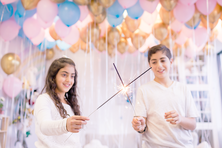 A girl and boy with sparklers. Dozens of blue, gold, and pink helium balloons float overhead.