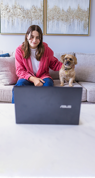 A teenage girl seated on a sofa, with a puppy by her side, looking into a laptop.