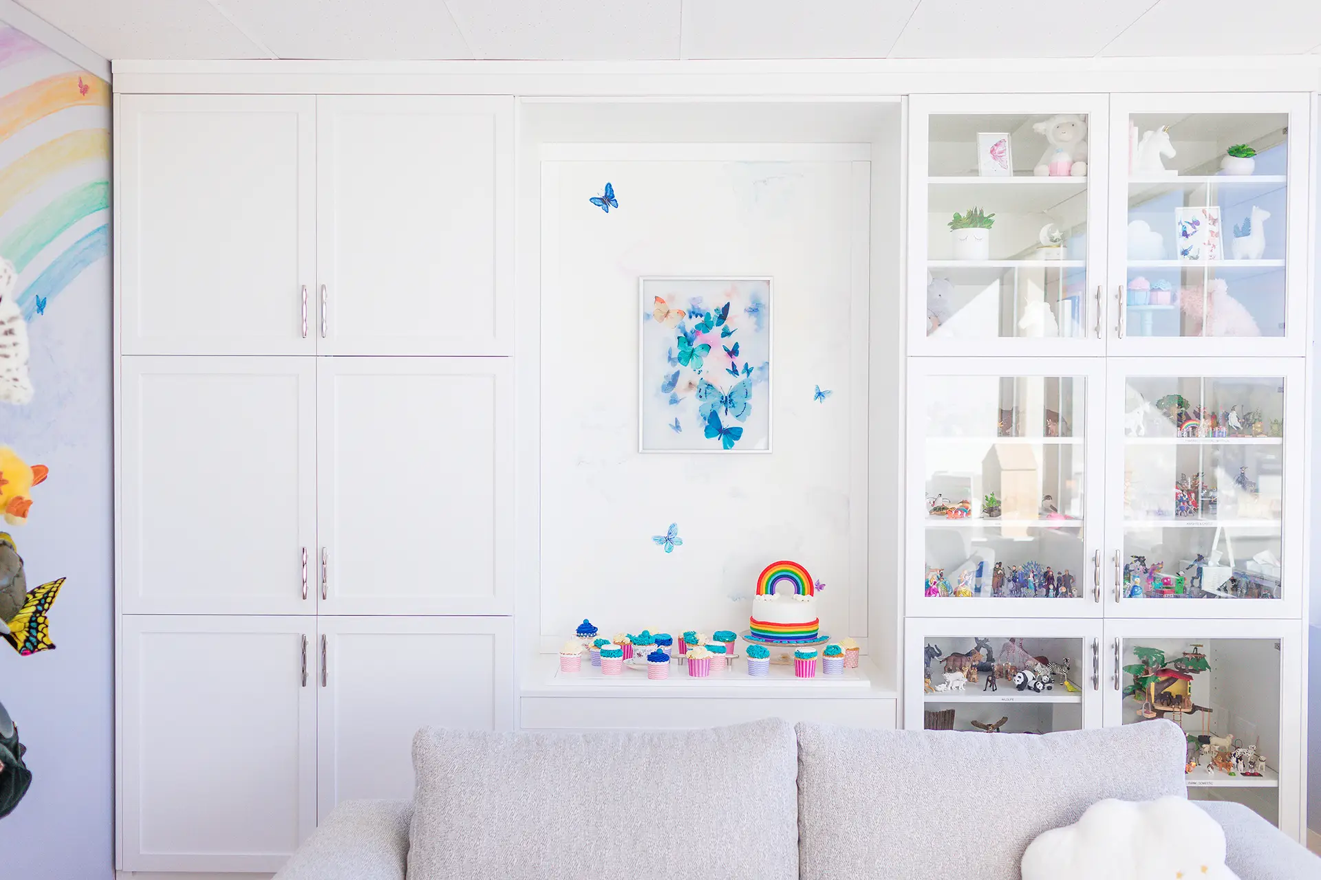 A soft couch in the foreground, with cloud shaped pillow. A butterfly painting on the wall, a rainbow mural on the left with toy puppets on a perch, and a toy cabinet filled with toys on the right.