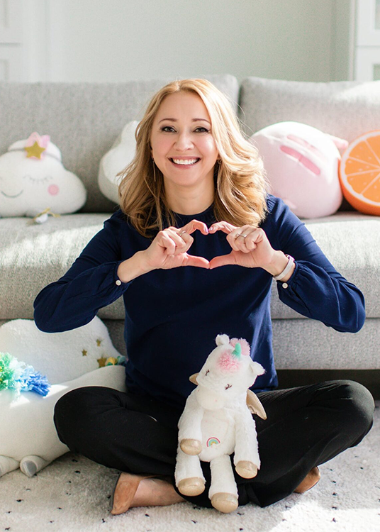 A child therapist sitting cross-legged on a white rug, making a heart shape with her hands. She is surrounded by fluffy toys