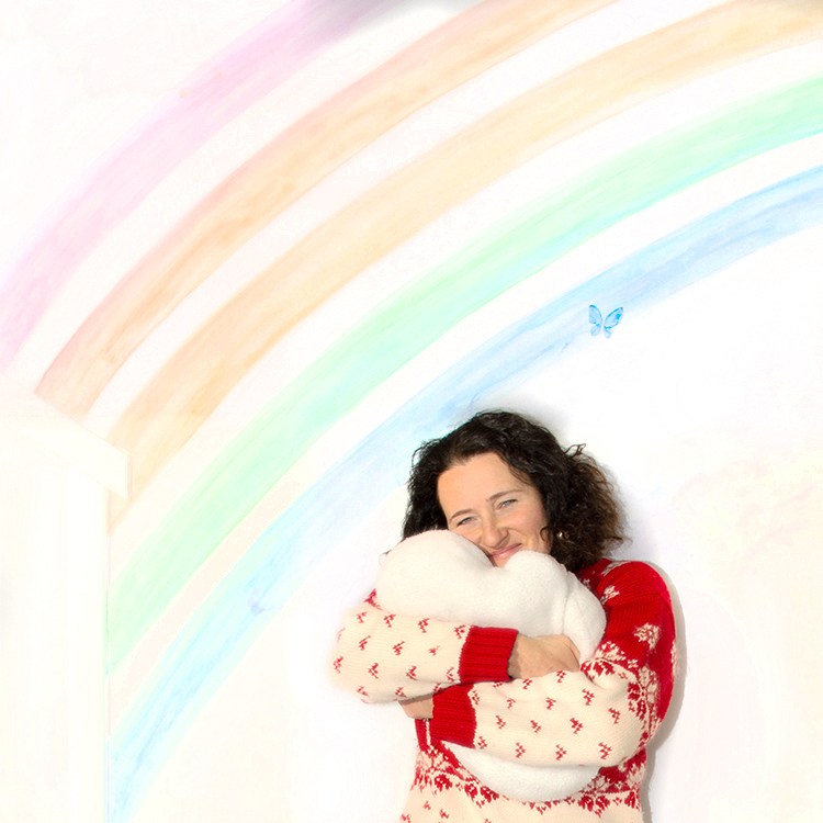 Nadia Ivanchikov, practicum student child therapist at Kids Reconnect, hugging a fluffy heart pillow, under a rainbow wall