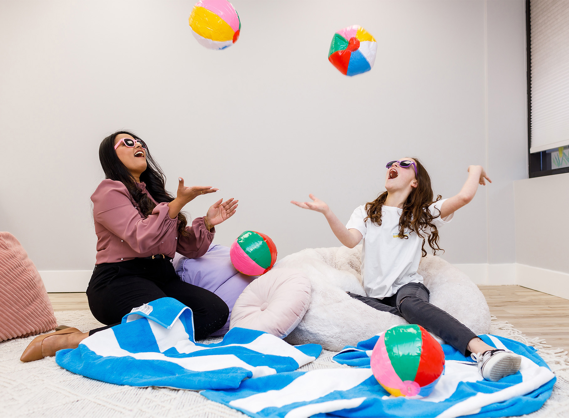 A child and her therapist wearing sunglasses, sitting on beach towels and throwing four colourful beach balls in the air, during a play therapy session.