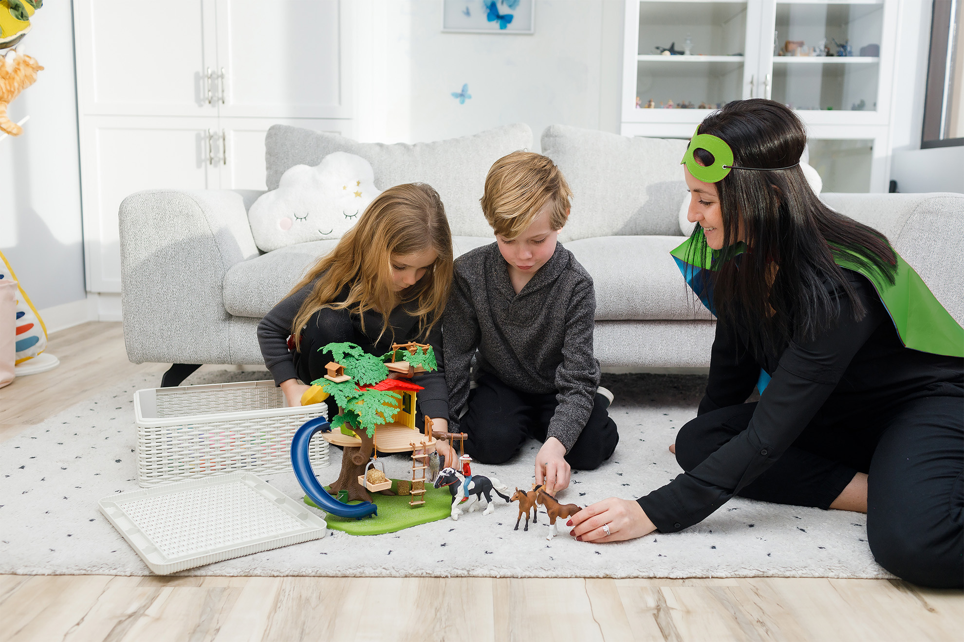 A child therapist wearing a Superhero cape in a play therapy session with two kids, a girl and a boy. They are sitting on a playroom rug, playing with a tree house toy and toy horse figures.
