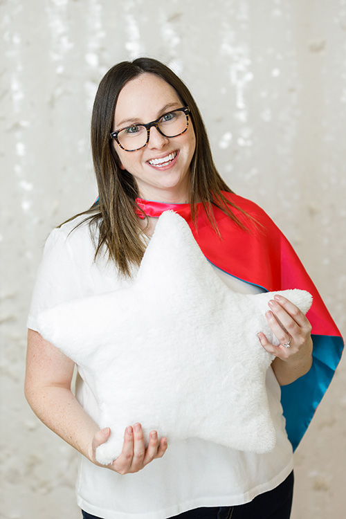 A smiling woman wearing a pink and blue cape holds up a white star-shaped fluffy toy stuffy. Jessica Donovan, Registered Social Worker at Kids Reconnect