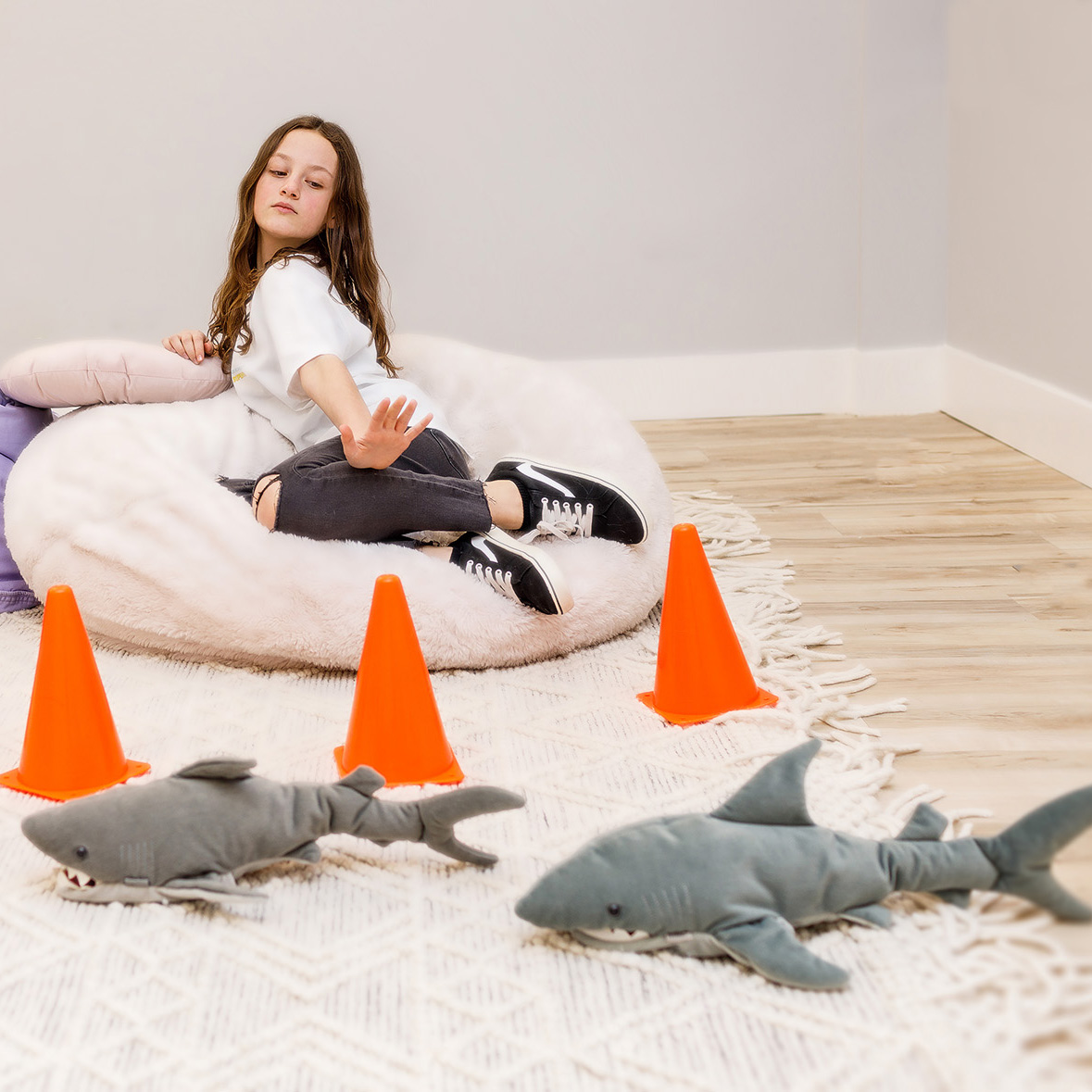A girl, seated on a pink pouffy cushion, holds her hand as a barrier. Toy traffic cones surround her, while two toy stuffy sharks swim around the play therapy rug.