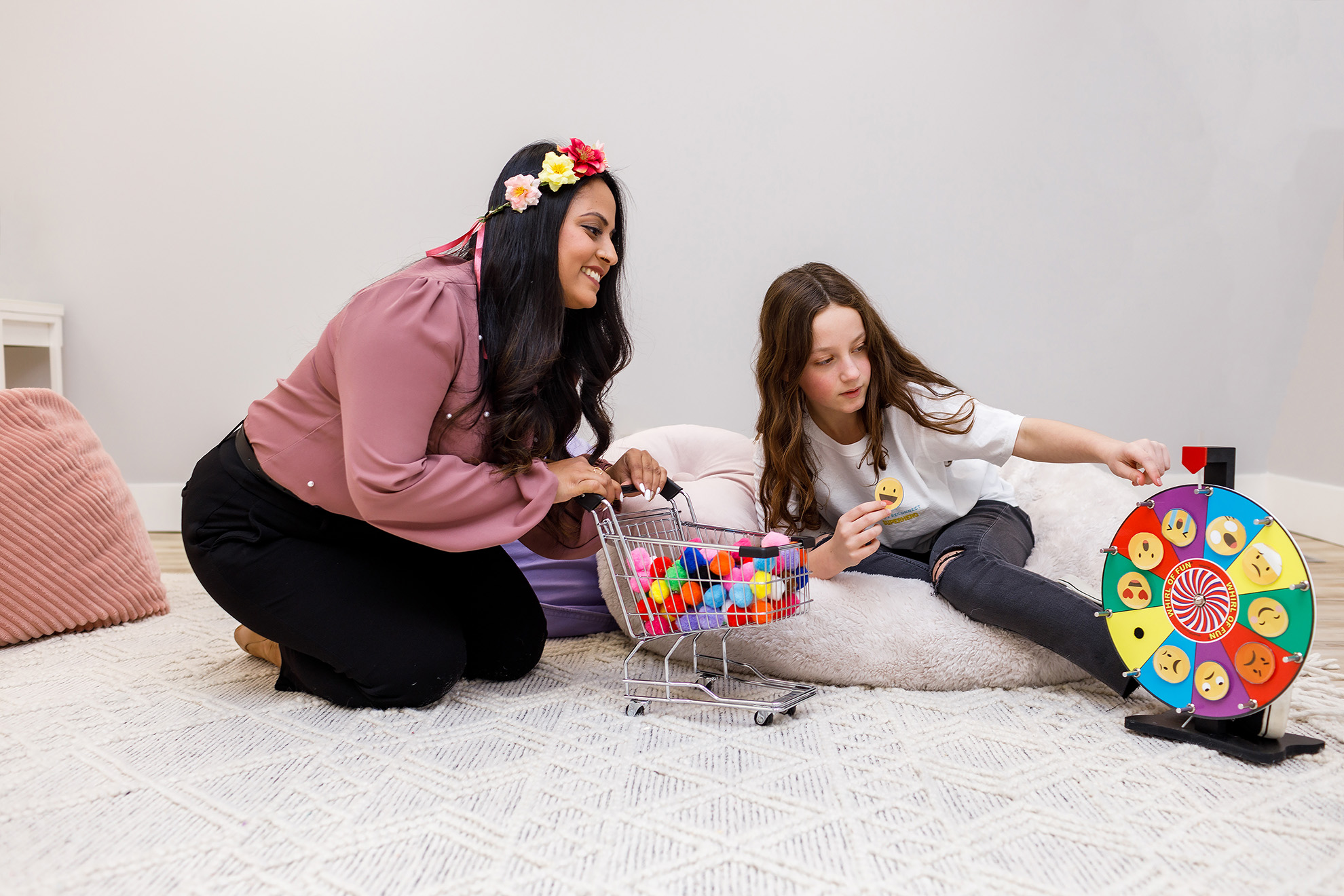 A child therapist and a child, sitting on a plush carpet. The therapist is holding a small toy shopping cart filled with coloured pompons; the young girl is spinning a wheel with different feelings emojis (smiling, love eyes, sad, etc.)