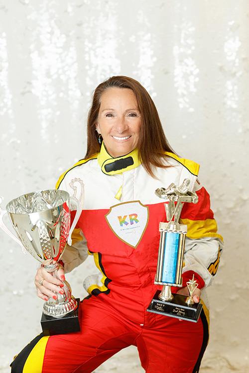 Headshot: Christine Boyd, MSW, RSW, child therapist at Kids Reconnect. Christine is wearing a car racing jumpsuit and holding two trophies, one in each hand.