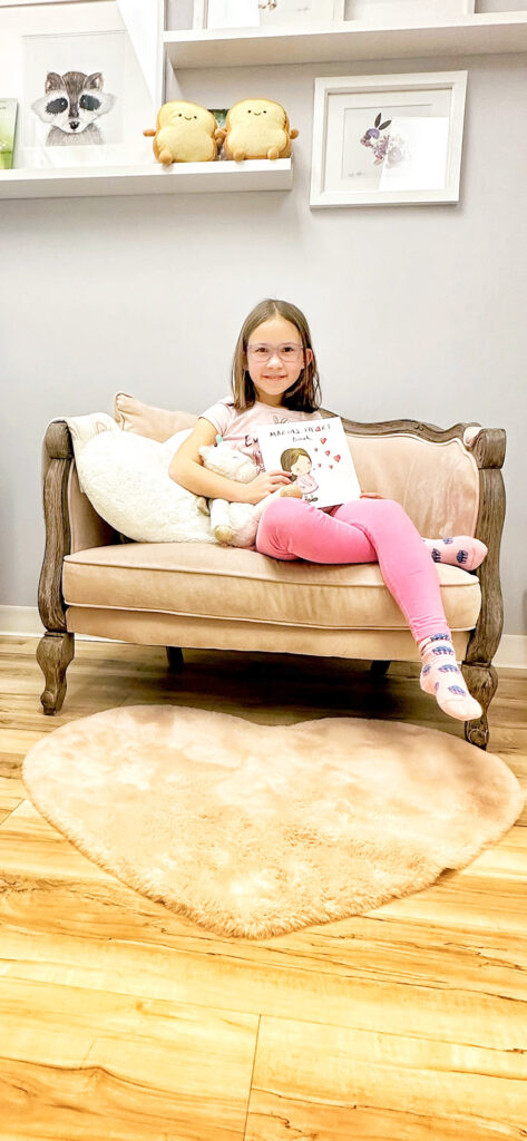 A little girl sitting in a child-size pink sofa holding a children's book called "Maria's Heart Book".