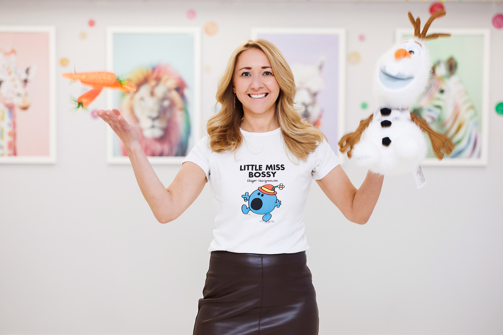 A play therapist smiles as she holds a toy snowman in one hand and juggles three toy carrots in the air.