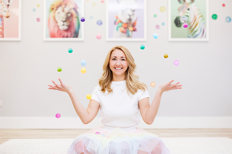 A child therapist sitting on the floor in a playroom, juggling pompoms in the air. Pictures of a colourful giraffe, lion, llama, and zebra in rainbow colours are in the background.