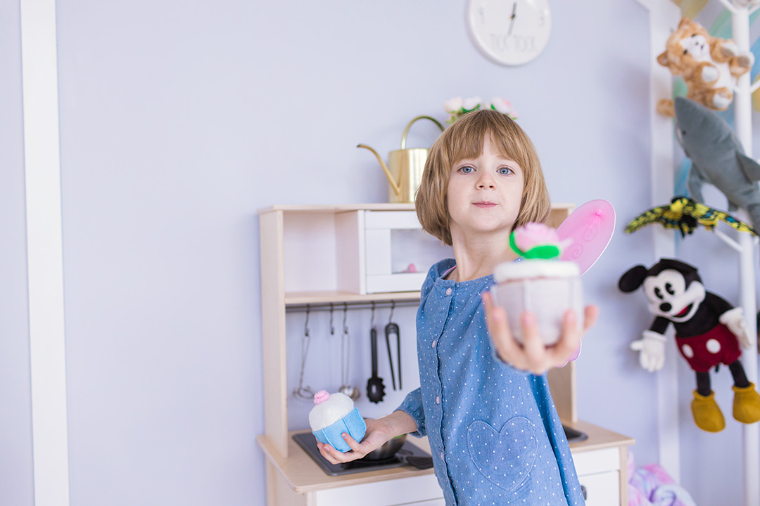 A child stands in a child therapy playroom. She smiles as she holds toy cupcakes, one in each hand. She is reaching out with her left hand, to share a cupcake.