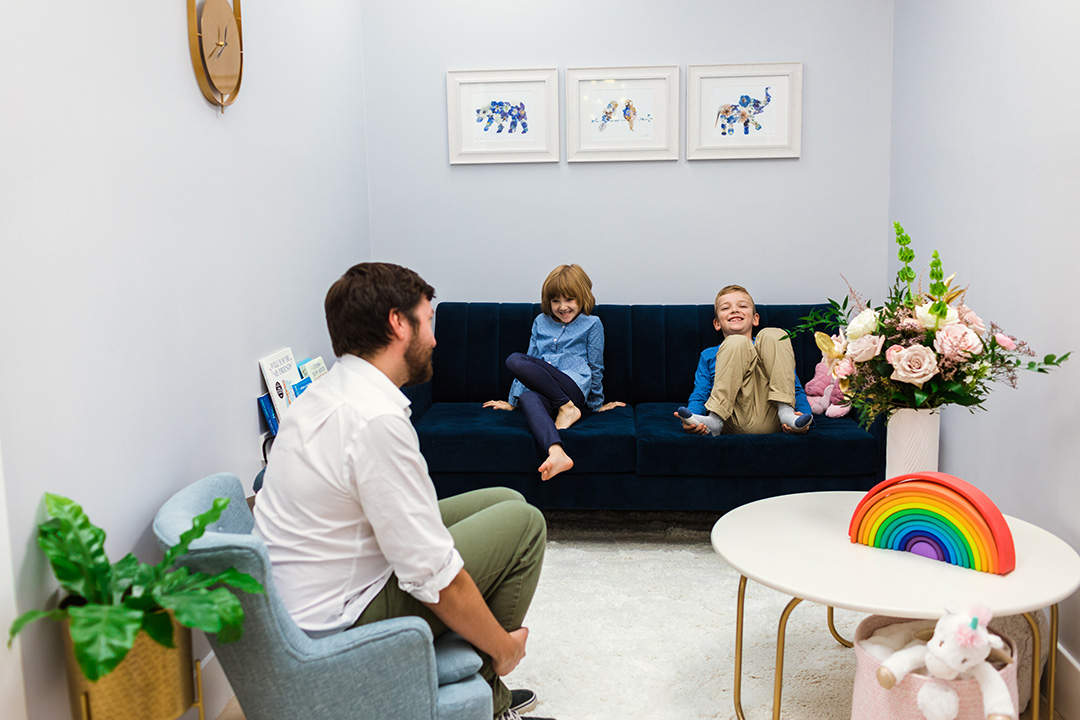 Two kids grin playfully at their father. The children are seated on a couch, facing their Dad, who is sitting on a small kid-size armchair.