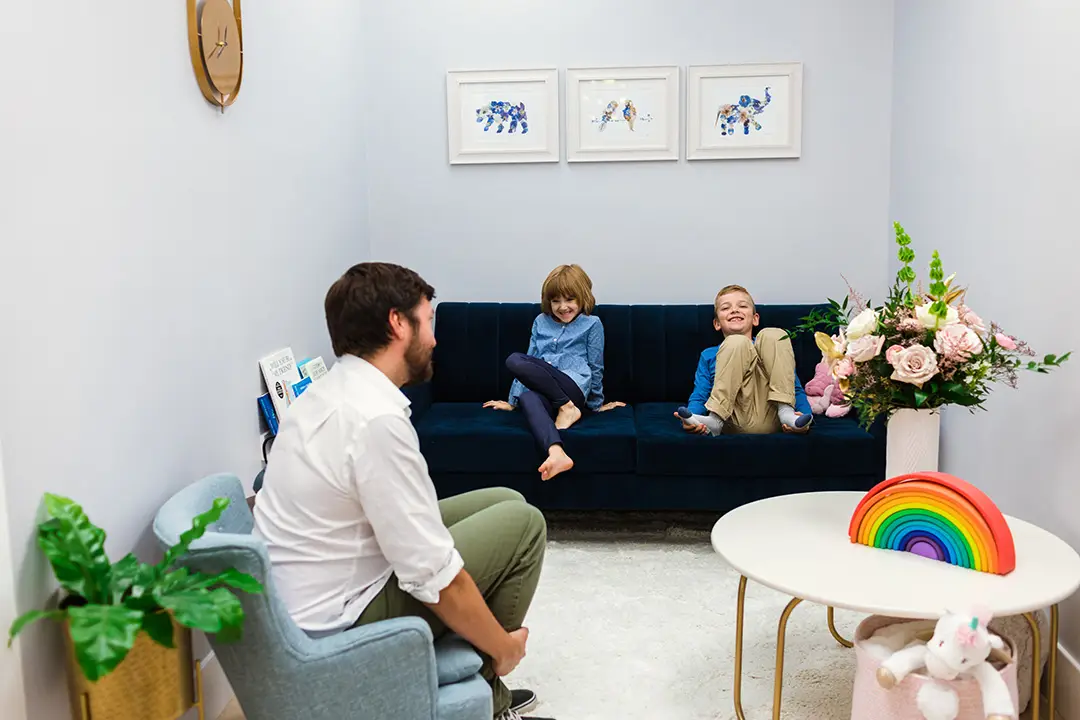 Two kids grin playfully at their father. The children are seated on a couch, facing their Dad, who is sitting on a very small kid-size armchair.