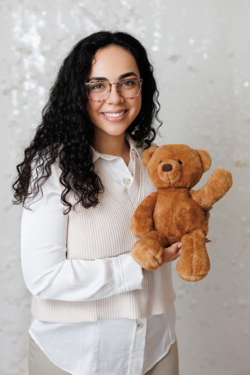 RIta Motta, MSW, RSW, child therapist at Kids Reconnect smiles as she holding a teddy bear who is waving his paw to say hi.