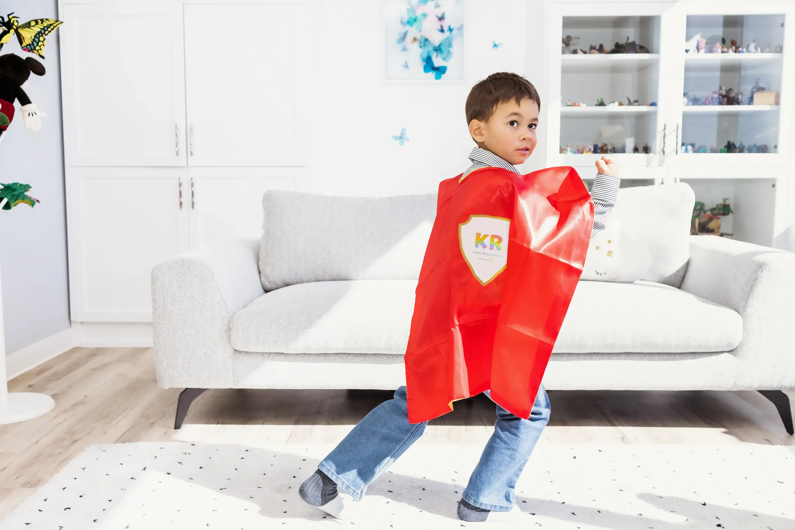 A boy stands in a power pose, making a fist with his right hand. He is in a playroom, wearing a Superhero cape with the Kids Reconnect logo.