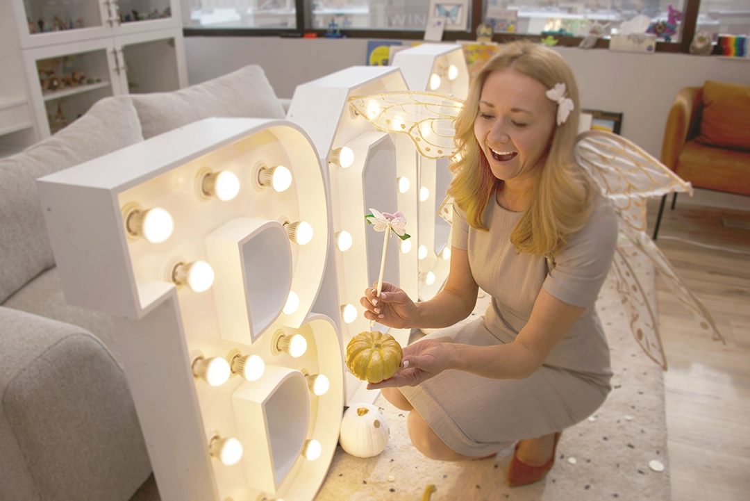 A woman wearing fairy wings sitting down in front of a large illuminated sign that says "BOO", as she holds a magic wand over a gold pumkin.