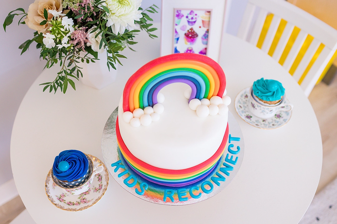 A beautiful cake on a child-sized table. The cake is decorated with rainbow icing. It is placed on top of a silver plate decorated with fondant letters that spell "KIDS RECONNECT". Also on the table, there are two teacups with icing decorated cupcakes, a vase with fresh flowers, and a framed watercolour illustration of cupcakes.