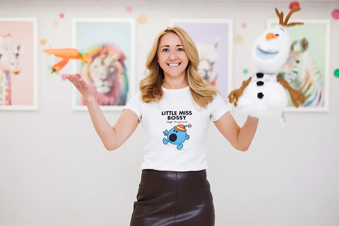 A play therapist smiles as she holds a toy snowman in one hand and juggles three toy carrots in the air. In the background, a picture of a rainbow-striped zebra, a rainbow spotted giraffe, and a rainbow-maned lion - playful imaginary creatures that spark kids' joy of play to grow and thrive.