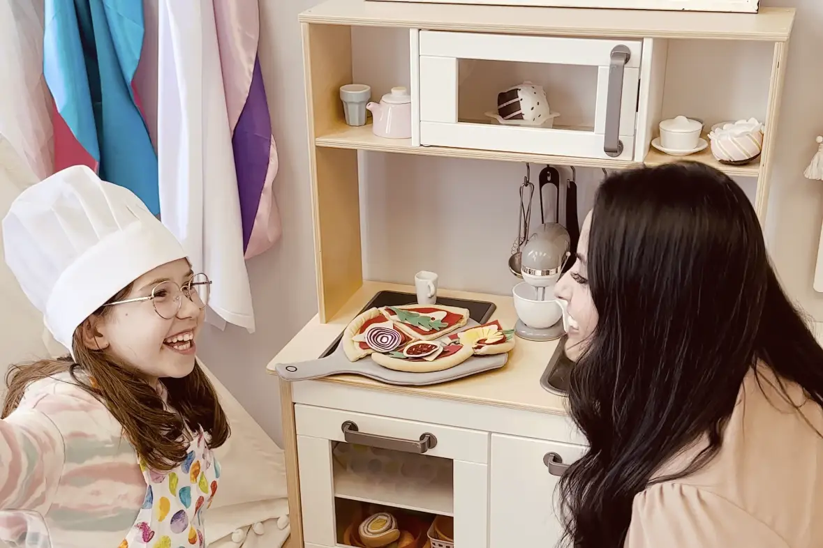 A girl wearing an apron and a chef's hat smiles with her therapist. There is a toy kitchen in the background.