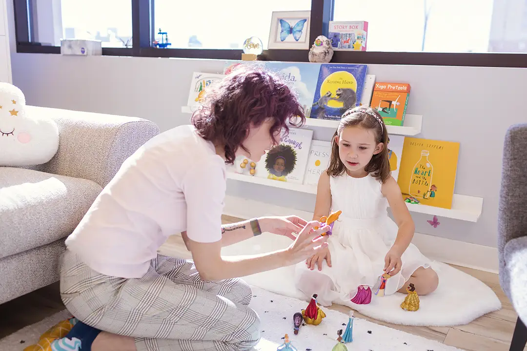 A child and a therapist playing with princesses and superhero figurines in a play therapy room. They are sitting on the carpet.