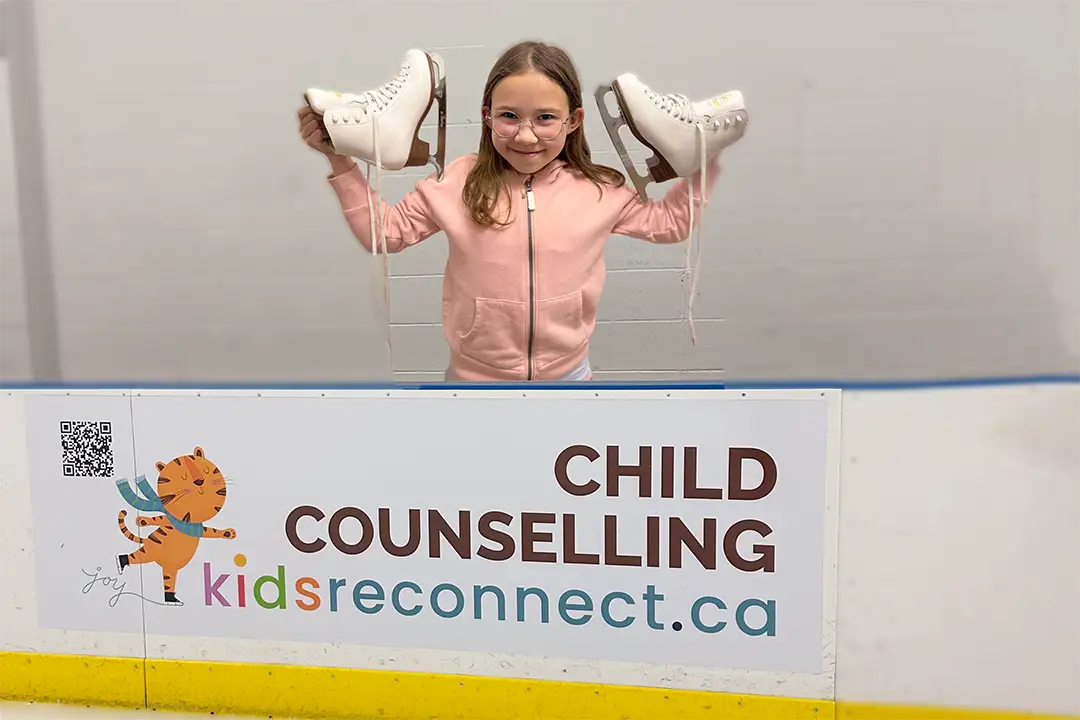 A girl smiles as she holds up her figure skates as she stands behind a rink board in an arena in Calgary. The rink board advertising sign features a cartoon tiger ice skating and says: CHILD COUNSELLING, kidsreconnect.ca "