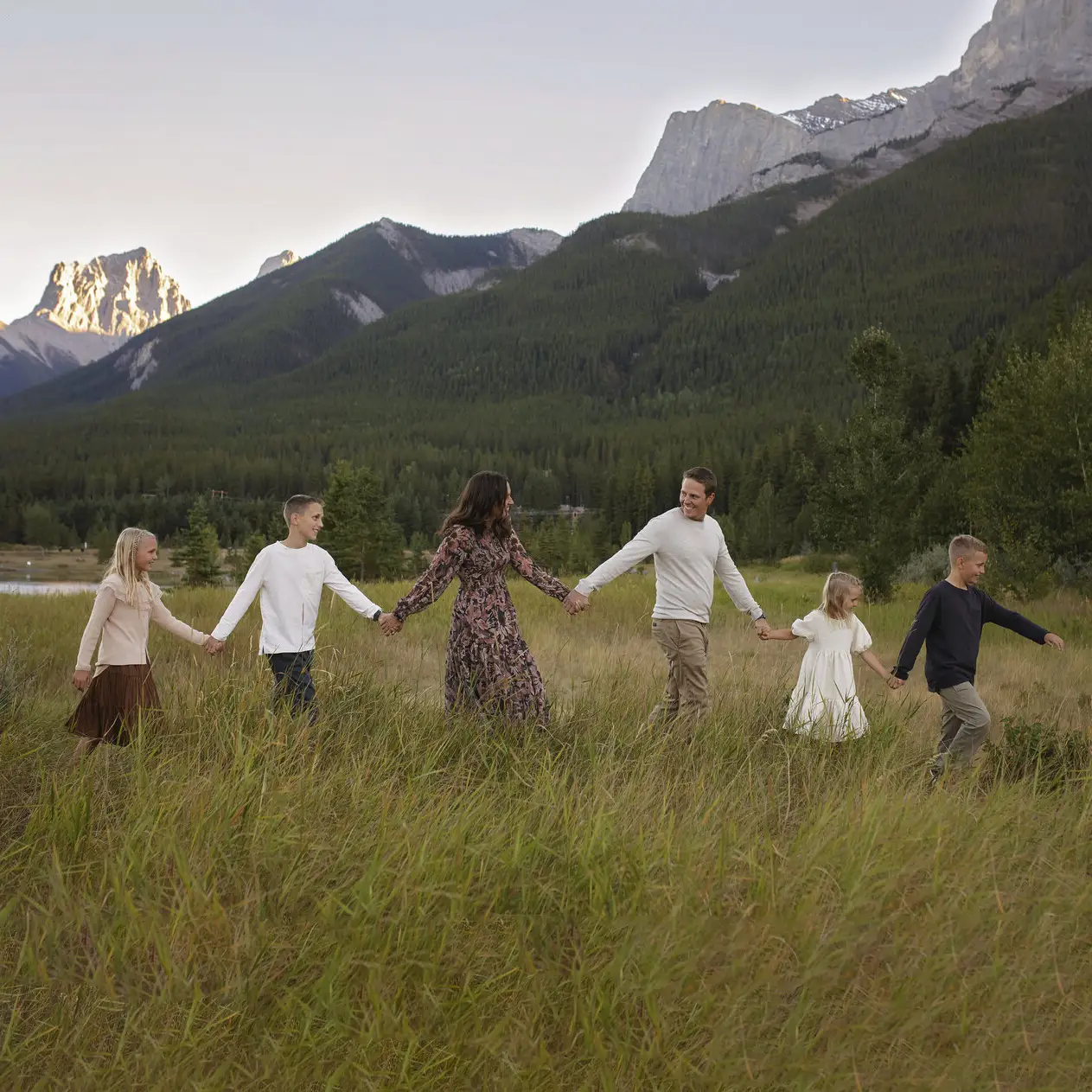 A family portrait with the Rocky Mountains in the background. From left to right, a girl, a boy, a mother, a father, a girl, and a boy hold hands as they walk across a grassy field.