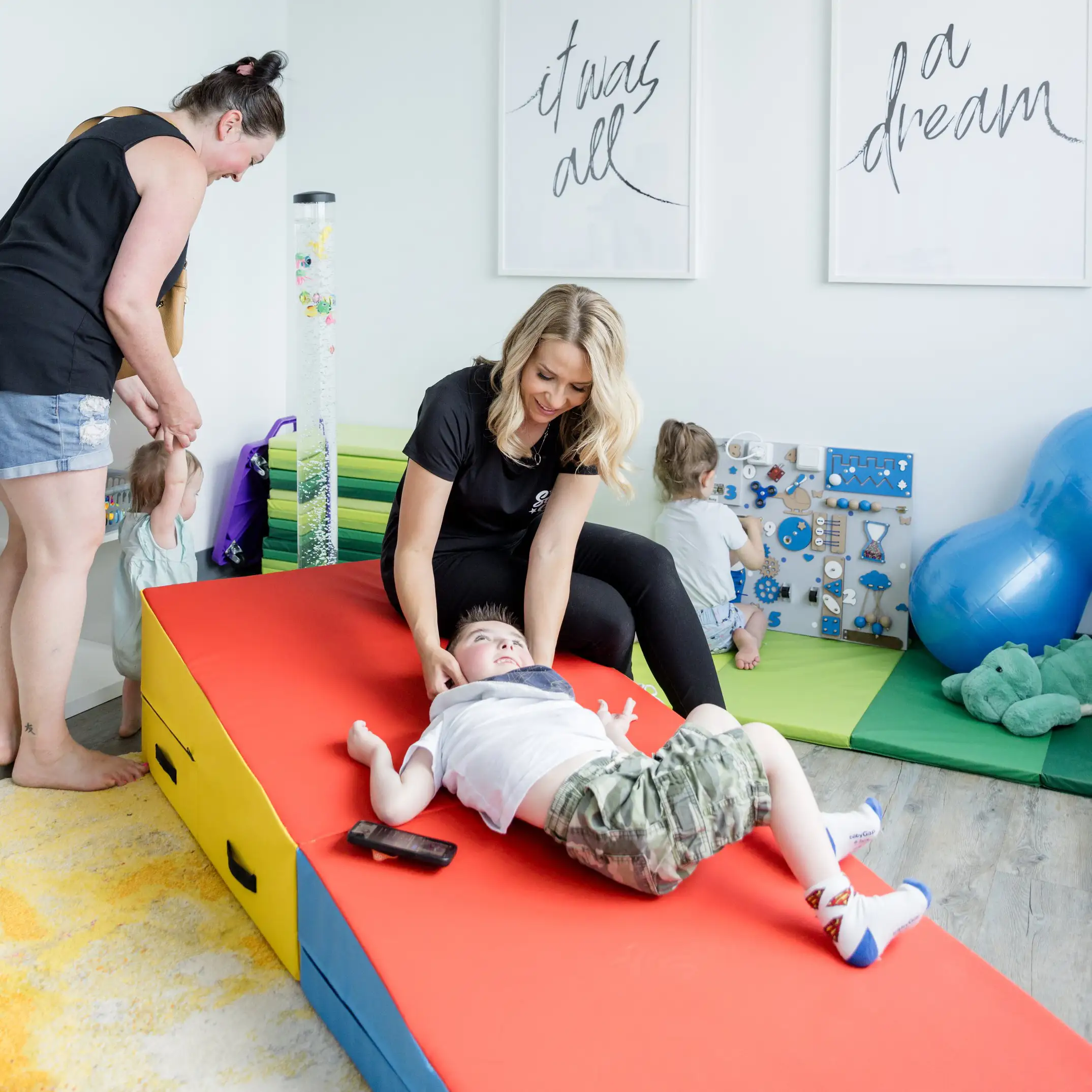 A pediatric massage therapist works with a child at Same Stars Wellness in Calgary. In the background, a therapist walks with a child, while a toddler plays.