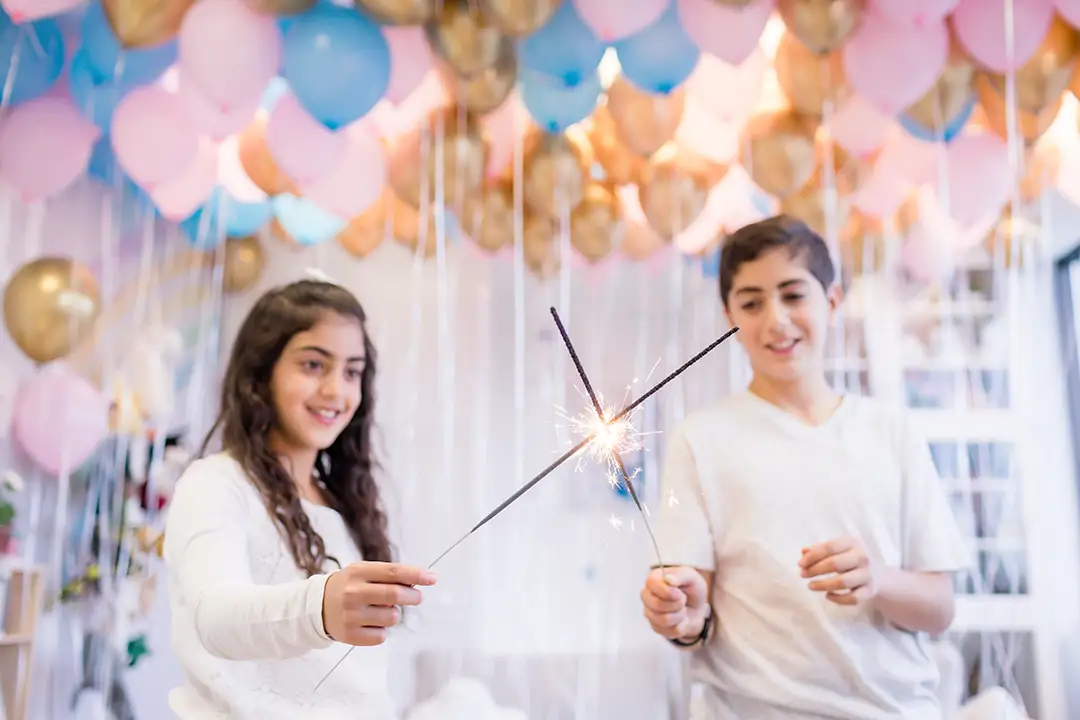 A girl and a boy smile as they each hold a sparkler. In the background, dozens of balloons float above them.