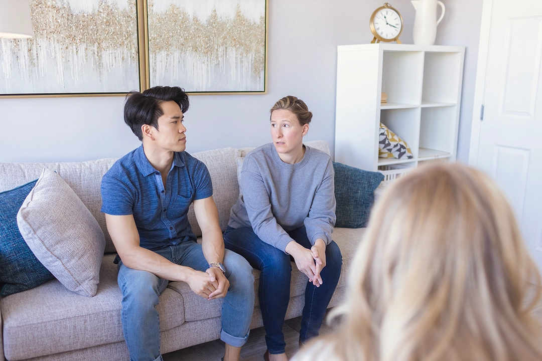 A father and mother sitting side by side on a therapy couch during a parenting session. The back of a therapist's head is in the foreground.