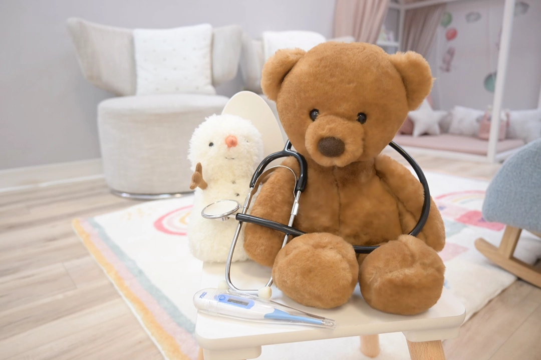 A teddy bear stuffy toy wearing a toy stethoscope and a stuffed toy snowman are sitting on a rug in a play therapy playroom. A thermometer is in the foreground.