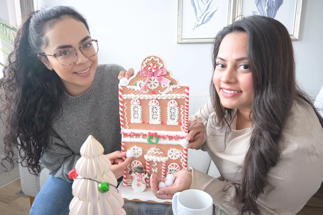 Two women smile at the camera as they build a gingerbread house.