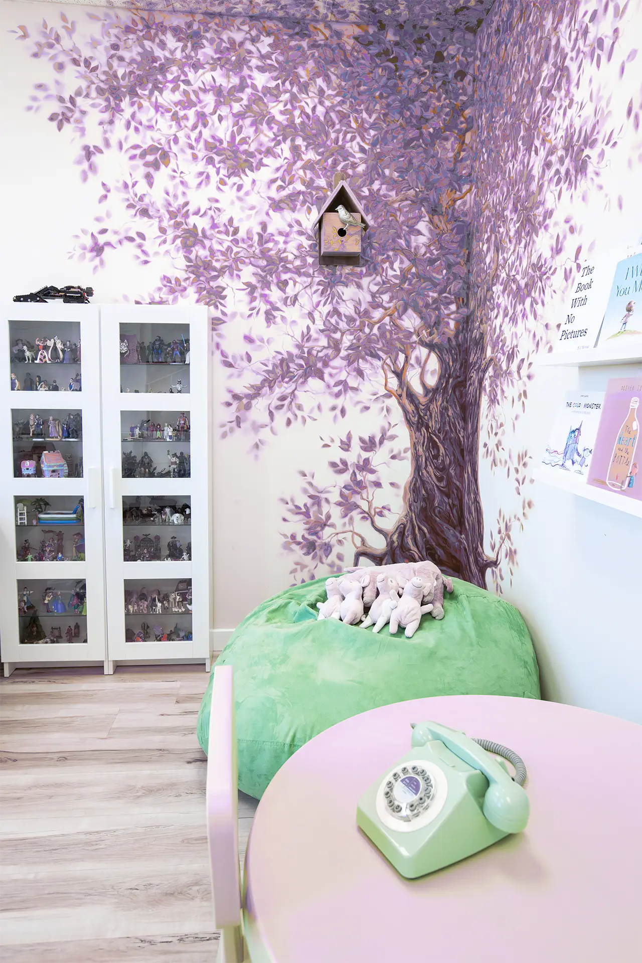 A playroom with a purple tree mural, toy piggies with their Mom on a green floor pillow, and a pink table with a green vintage phone in the foreground.