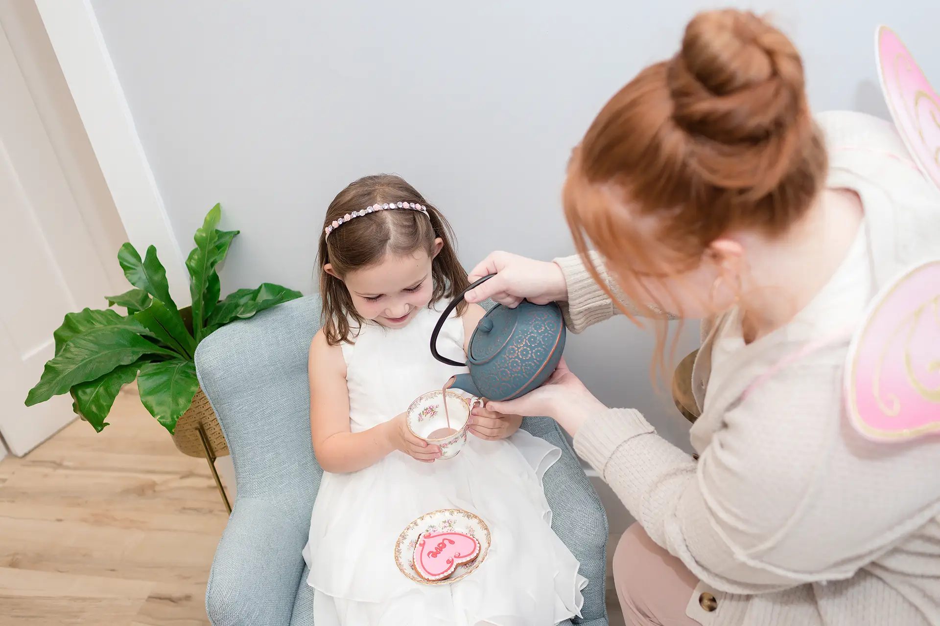 A girl, sitting on a child-size chair, holds a small cup and saucer as a woman wearing butterfly wings pours tea from a teapot. The child also has a plate with a heart-shaped cookie that says "love".