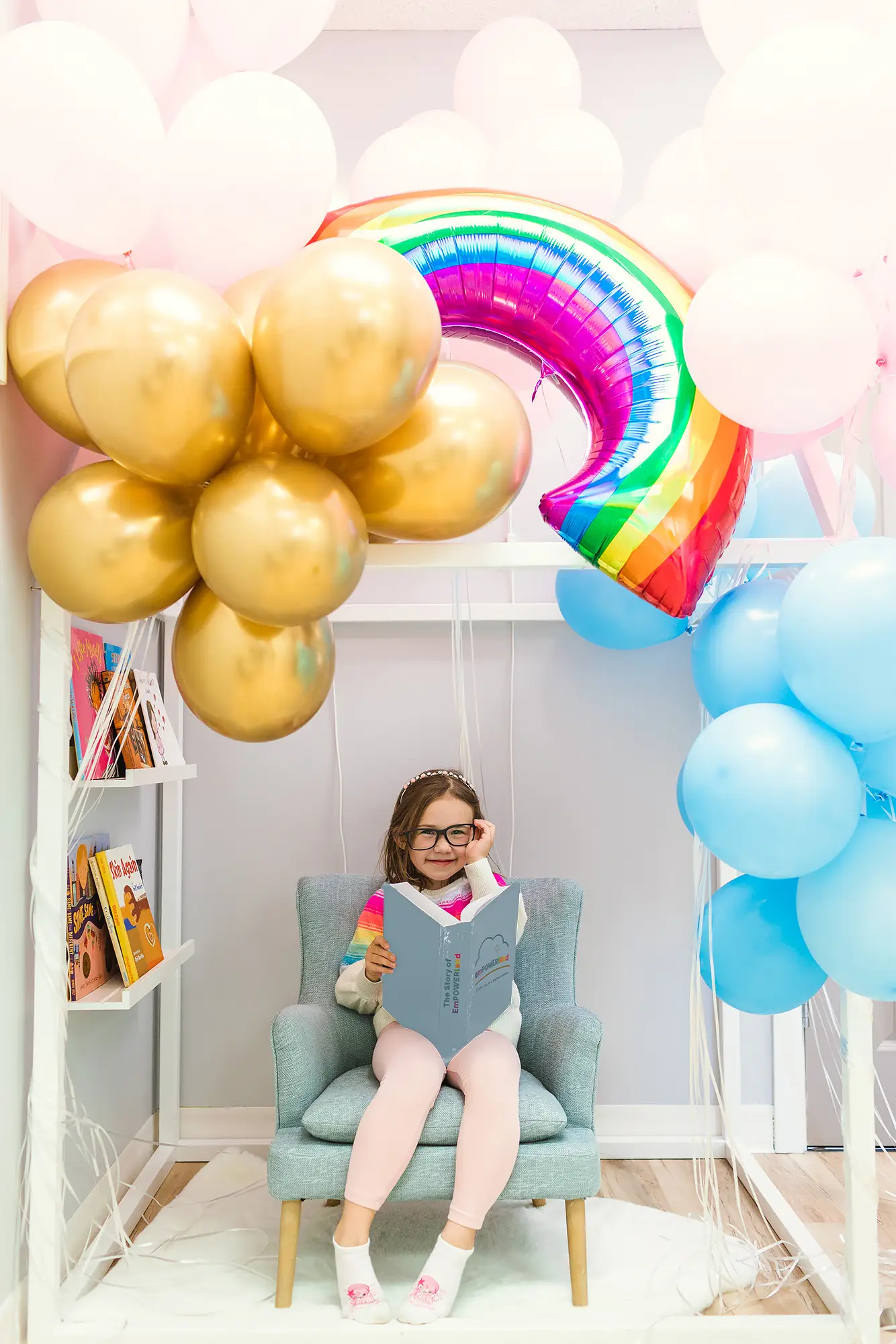 A child smiles as she reads The Story of EmPOWERland. She is wearing toy glasses. Gold, pink, blue, and rainbow balloons shine overhead. She is sitting on a child-sized chair in a play house.