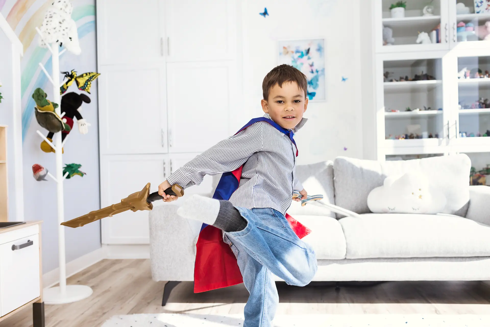 A child, wearing a red and blue cape, brandishes two plastic sword, one in each hand, while skillfully performing a turnkick. He is in a child counselling playroom at Kids Reconnect.