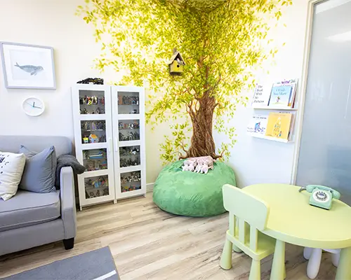 A painted tree mural in a playroom at Kids Reconnect. A sofa, a pouffy sea, a child-sized table and chairs, and a cupboard full of toys and some books are all part of a fun playroom especially designed for kids!