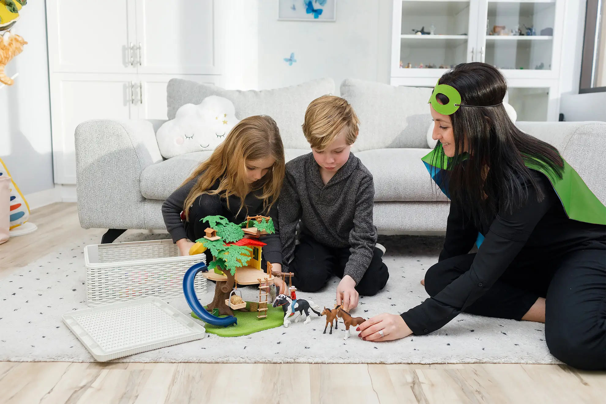 A child counsellor wearing a Superhero cape in a play therapy session with two kids, a girl and a boy. They are sitting on a playroom rug, playing with a tree house toy and toy horse figures.