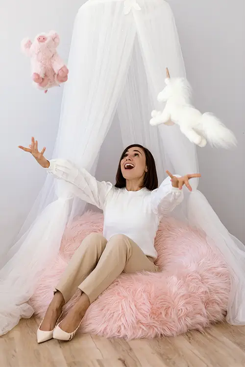 A child psychologist (Marcella Galizia) is sitting on a fluffy pink floor pillow, playfully throwing a flying toy piggy and unicorn in a playroom.