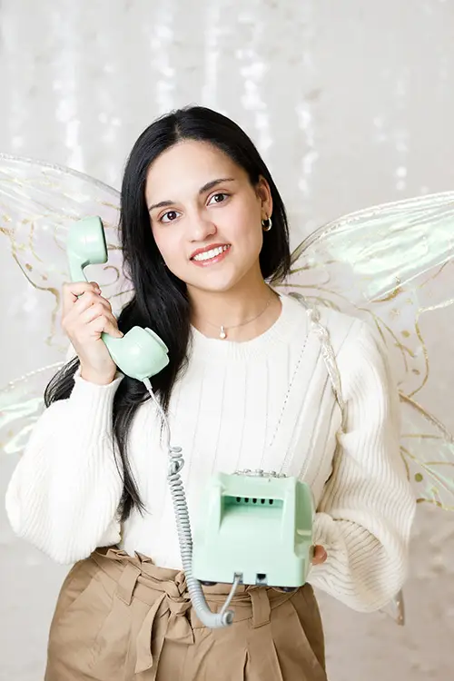 A woman wearing butterfly wings playfully holds a toy rotary phone. This is Mishaal Jannat's playful headshot.