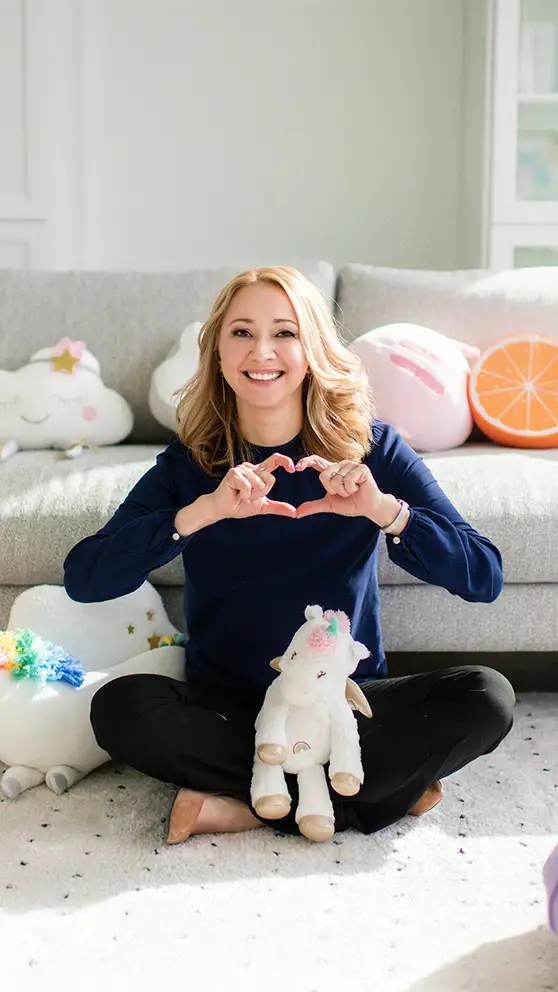 A child psychologist (Natalie Bergman) sitting cross-legged on a rug in front of a sofa, surrounded by stuffies and pillows. She is making a heart shape with her hands.