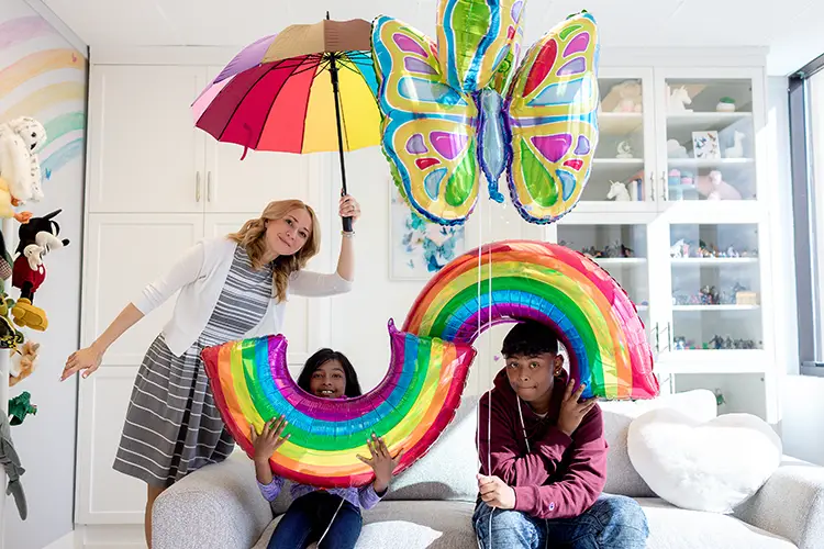 A child psychologist stands as she holds a rainbow coloured umbrella over two seated children. Each child is holding rainbow coloured balloons and a butterfly balloon. They are in a play therapy playroom.