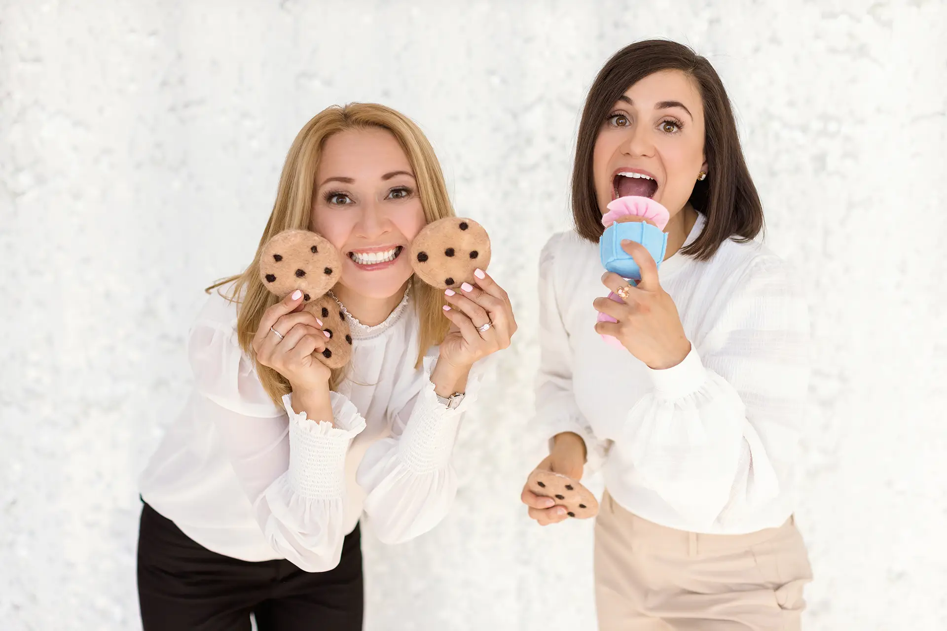 Two child psychologists, Natalie Bergman and Marcella Galizia, smile playfully. The woman on the left is holding two toy cookies; the woman on the right is pretending to eat a toy cupcake.